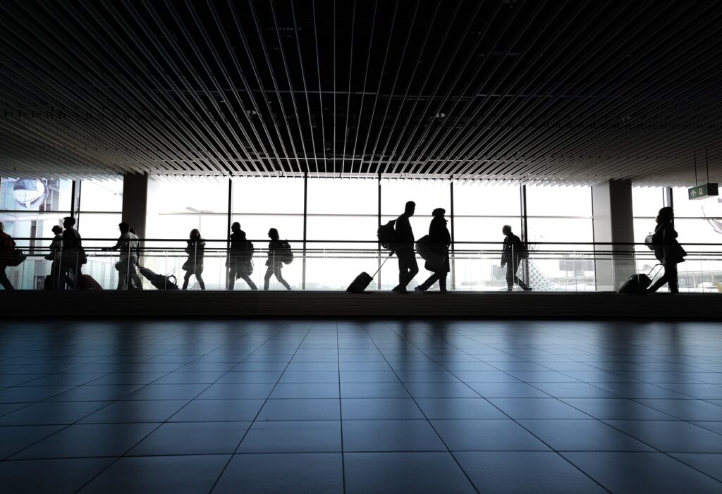 Voyageurs marchant dans un terminal d'aéroport avec des valises, silhouettés contre une grande baie vitrée.